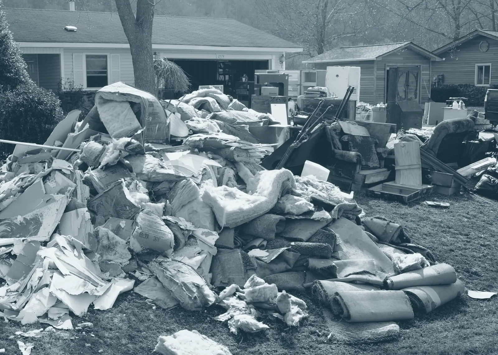 A large pile of wet insulation, broken drywall and rolled carpet on a front lawn, with furniture beside a neighboring house